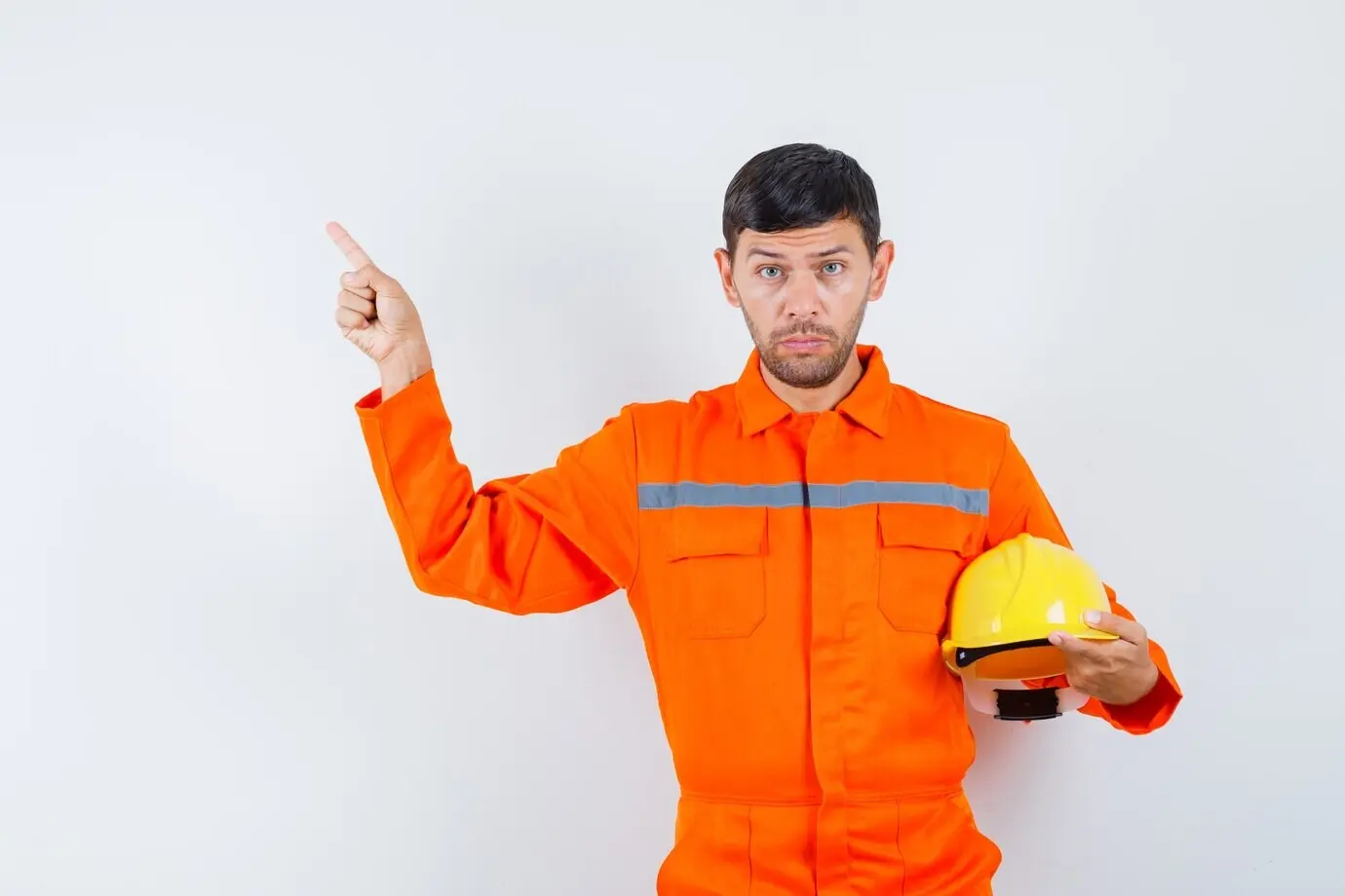 Ouvrier industriel tenant un casque, pointant vers le coin supérieur gauche, en uniforme, de face.